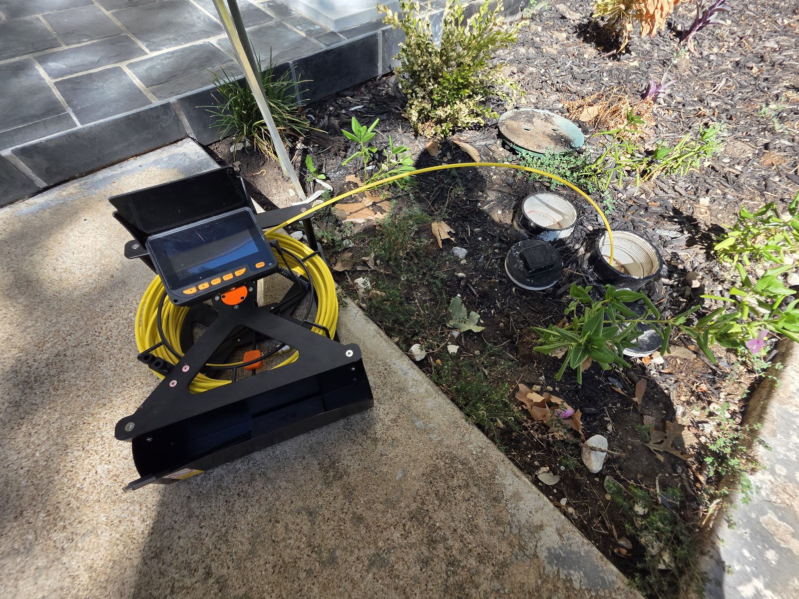 Carrete y monitor de la cámara de drenaje en una banqueta, con el cable amarillo de la cámara entrando a un registro abierto en una jardinera