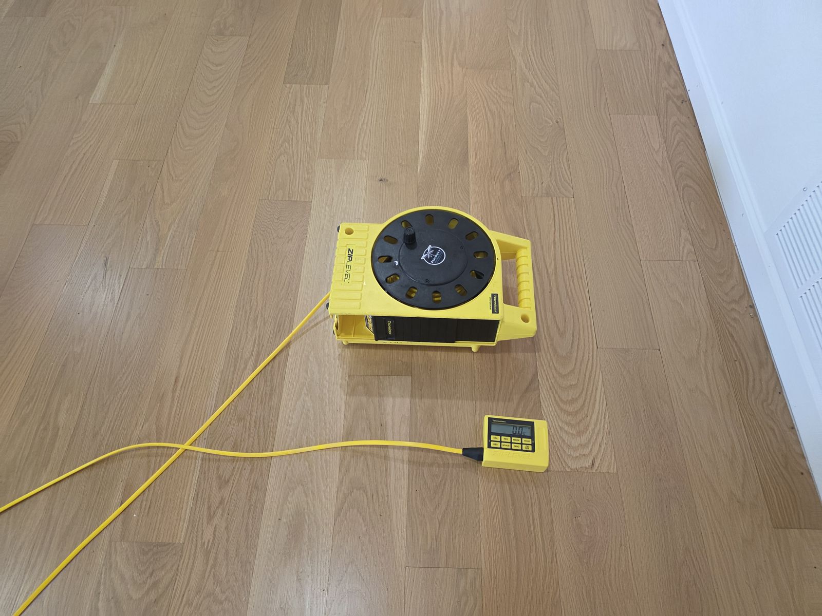 ZipLevel reference base and handheld readout connected by a yellow hose on a light oak floor